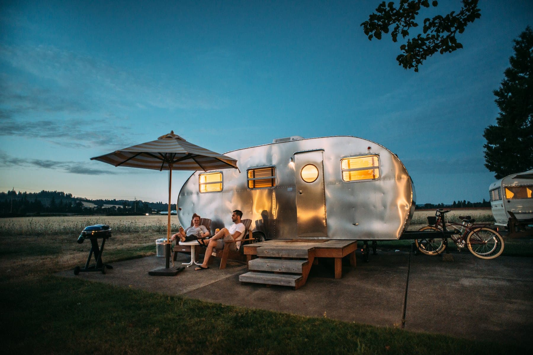 A couple seated in camp chairs in front of their RV trailer at dusk